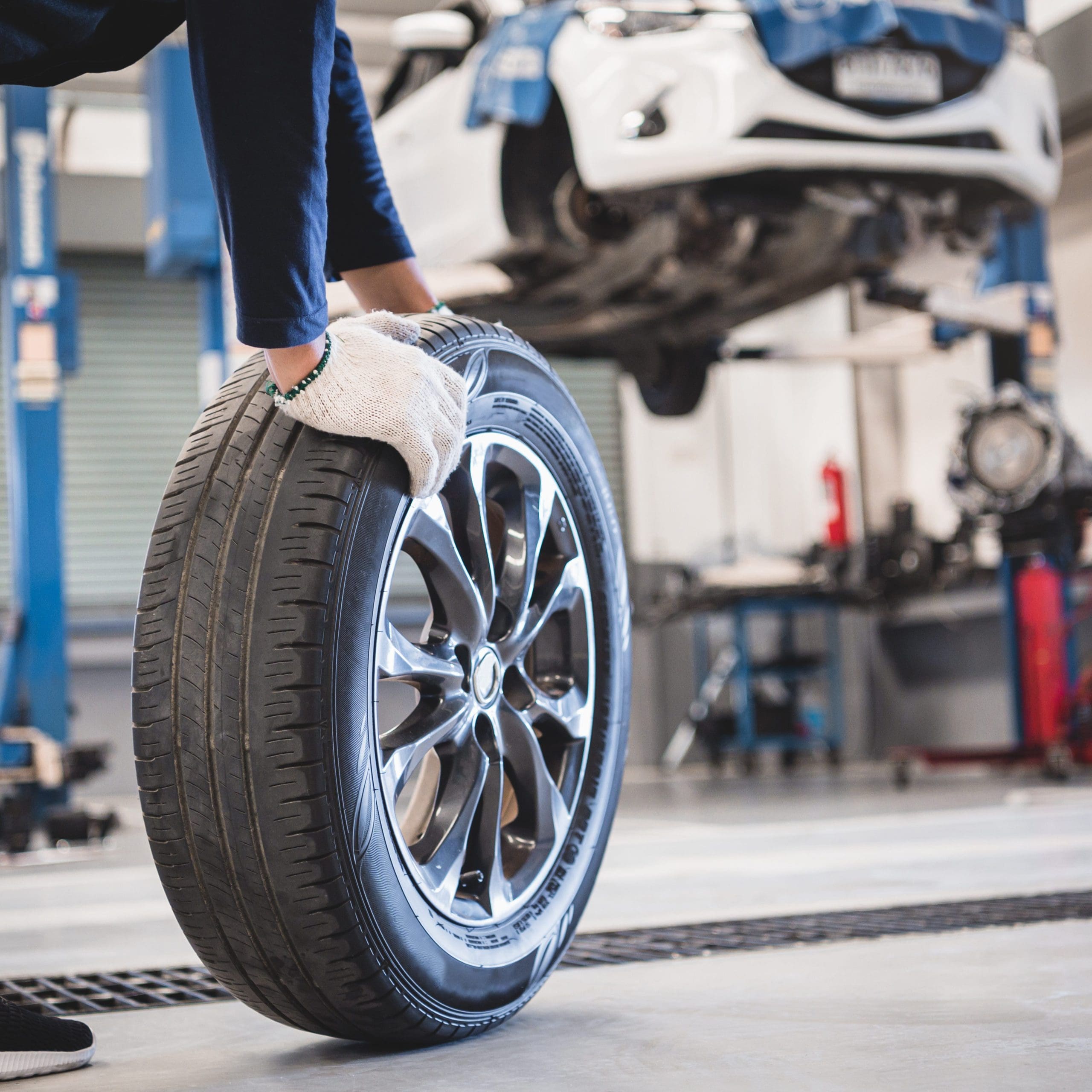 A person wearing gloves is handling a car tire in an auto repair shop during routine car servicing. A car is elevated on a lift in the background, and various tools and equipment are scattered around the garage, ready for anything from clutches to MOT testing.