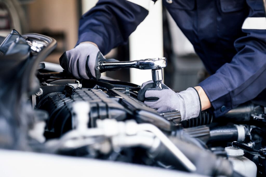 A garage mechanic in a blue uniform uses a socket wrench to work on a car engine. Engaged in their task, they don gray gloves as various engine components are visible, including hoses and parts of the engine block, all while ensuring thorough DPF cleaning.