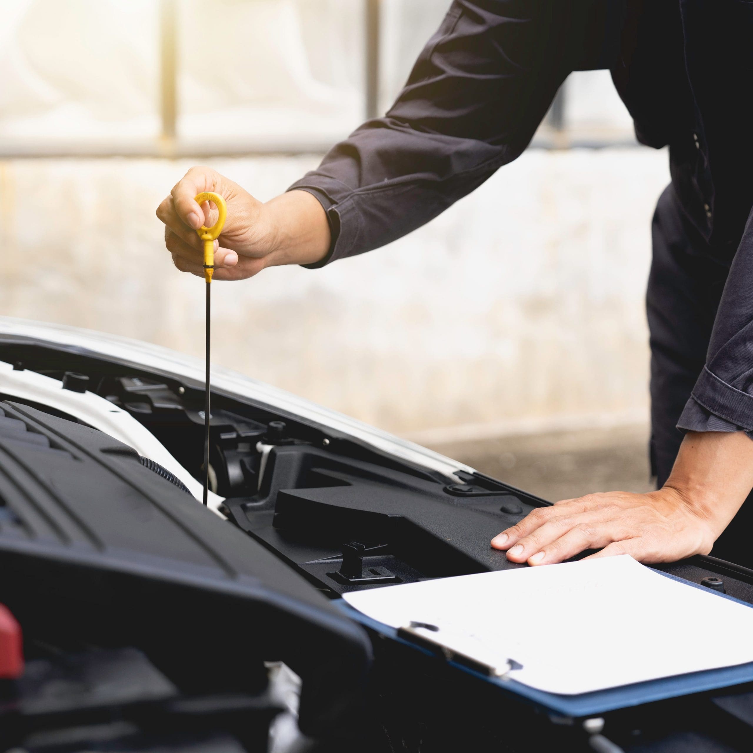A garage mechanic in a dark uniform checks the engine oil level with a dipstick. One hand holds the dipstick, while the other rests on the car's engine. A clipboard, possibly for an MOT testing checklist, is visible in the foreground on the car.