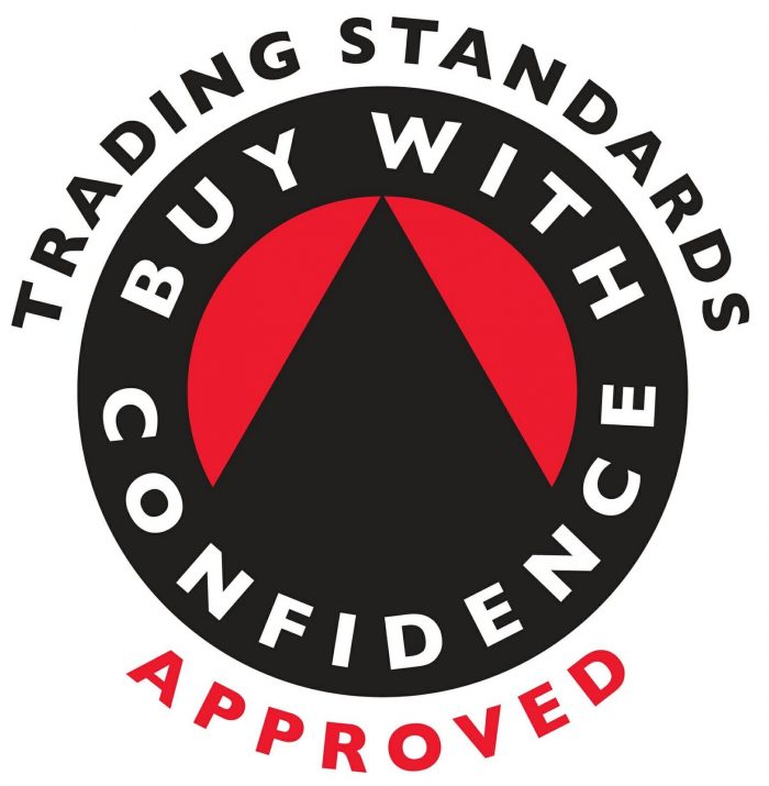 The image features a circular logo with "Trading Standards" at the top, "Buy With Confidence" in the center around a red triangle, and "Approved" at the bottom. The design, in white and red on a black background, conveys trust akin to crucial services like MOT Testing.
