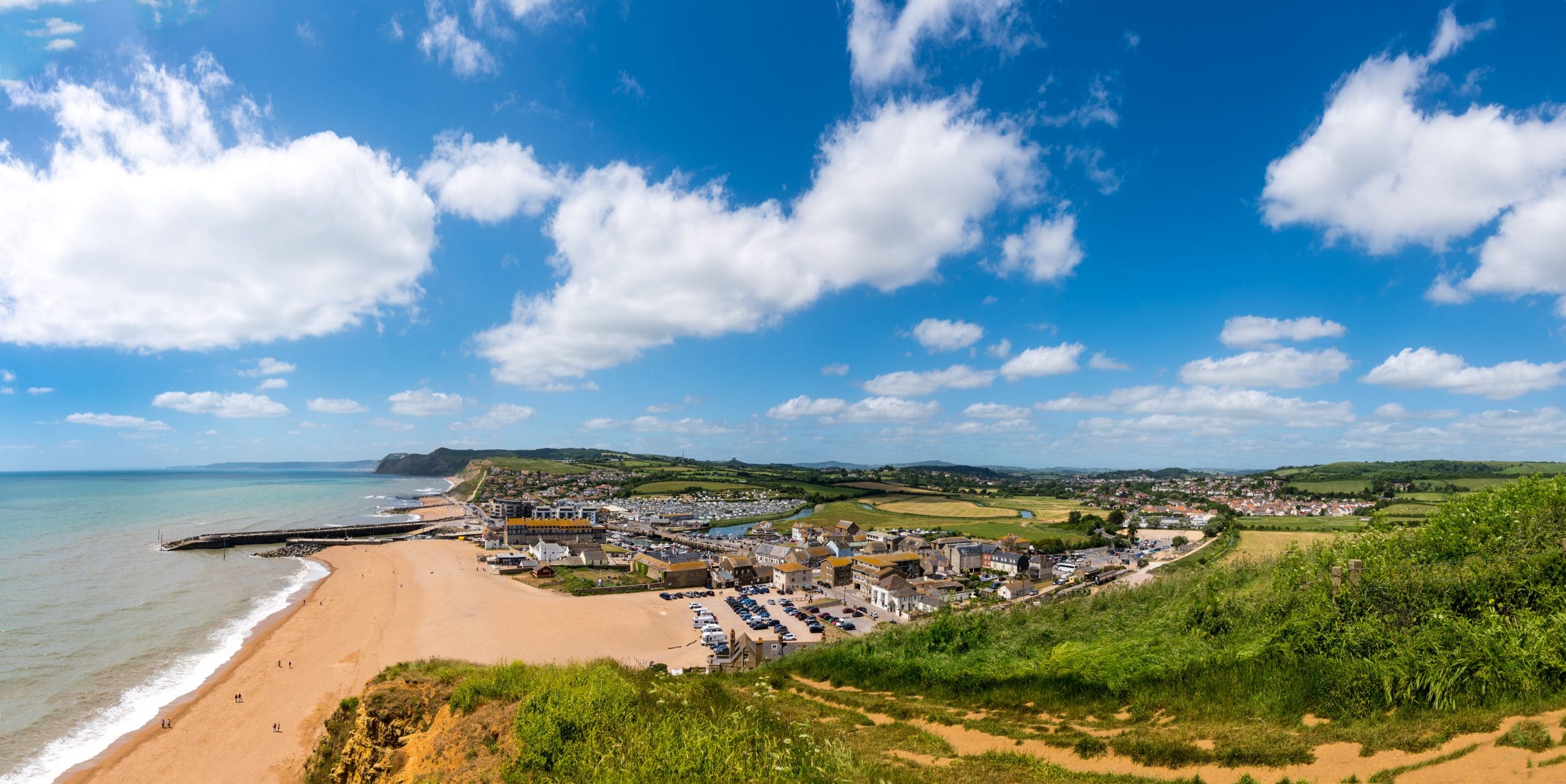 Panoramic view of a coastal town with a sandy beach and clear blue sky. The scene features grassy cliffs, a marina brimming with boats like clutches in motion, and green hills in the background. White clouds dot the sky, adding depth to the scenic landscape.