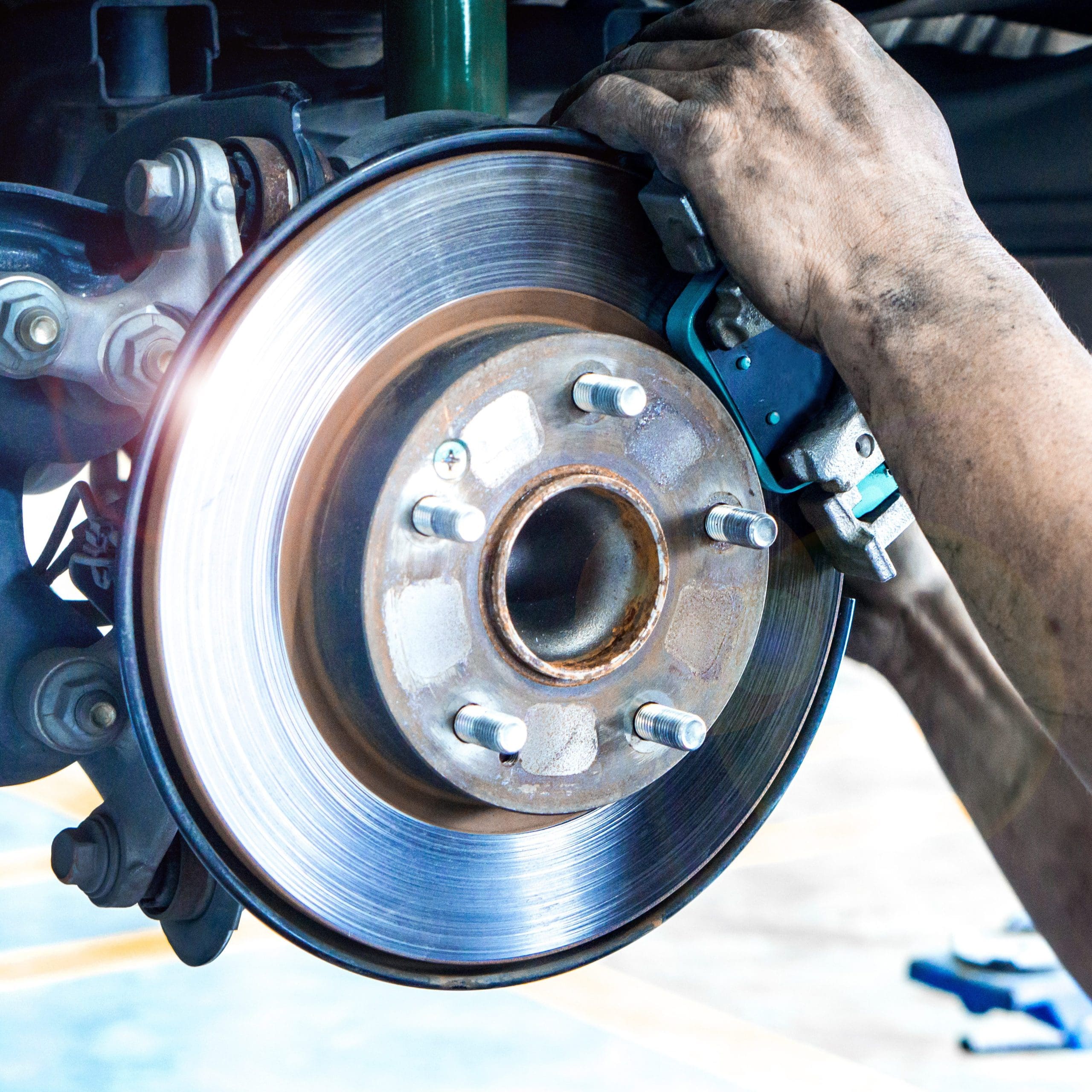 Mechanic's hands working on a shiny car brake disc and caliper during routine car servicing. The image highlights the metallic surface of the brake components and the intricate details of the braking system in a bustling workshop setting.