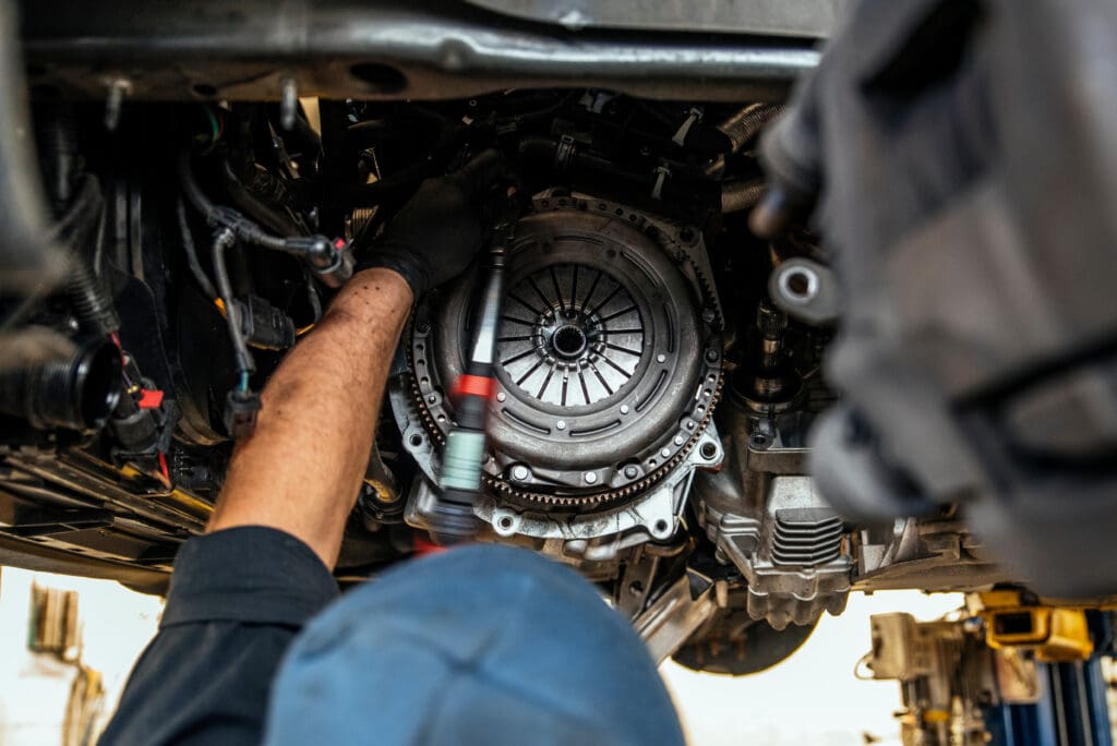 A mechanic diligently works under a vehicle in an automotive workshop, wielding tools to repair the clutch system. The focus is on the intricate clutch components, with glimpses of the mechanic's arms and head visible among the bustling environment of car servicing tasks.