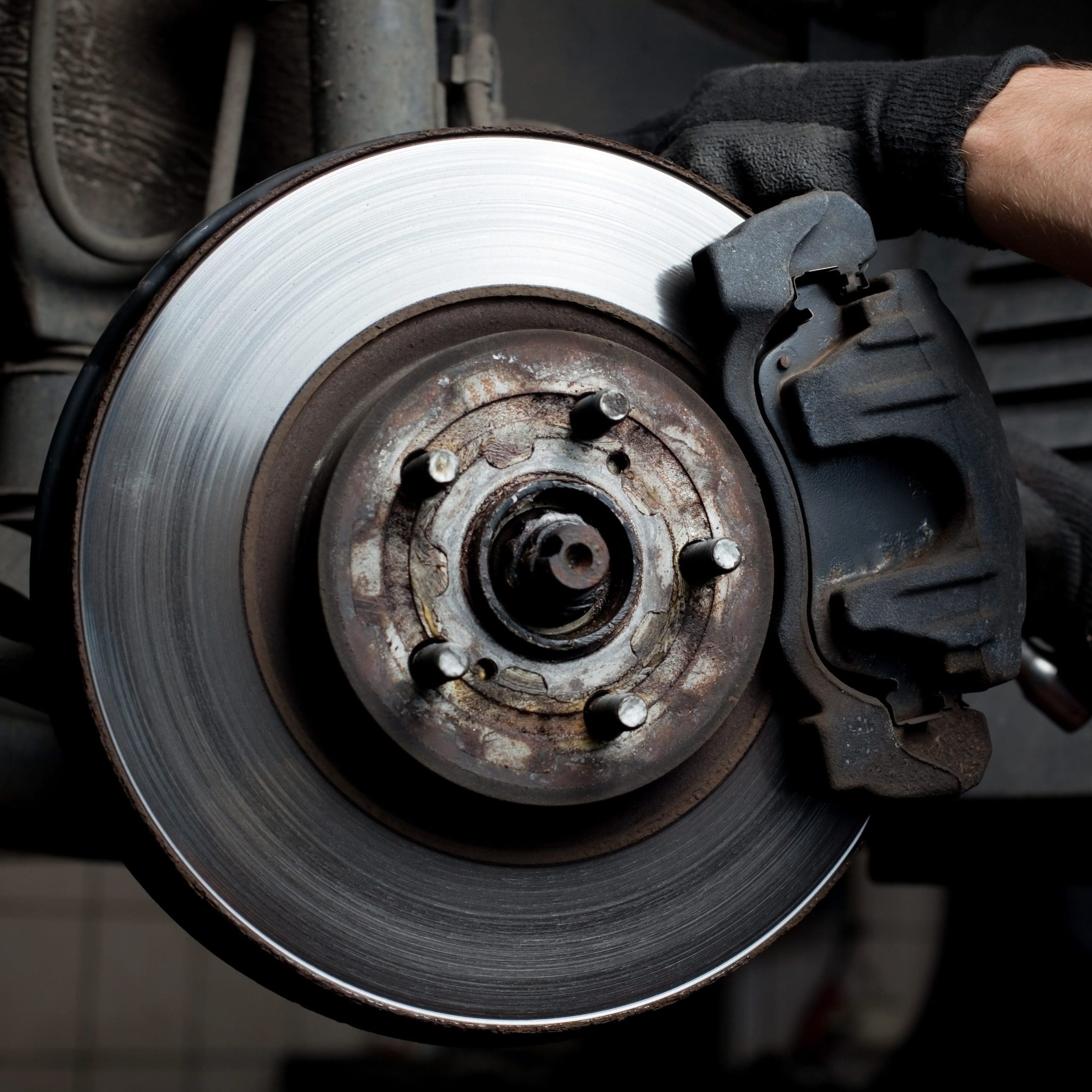 A gloved hand holds a car brake disc and caliper, showcasing the detailed mechanics familiar to any garage mechanic. The image focuses on the worn metal components, highlighting the circular disc's grooves and the rugged texture of the caliper.