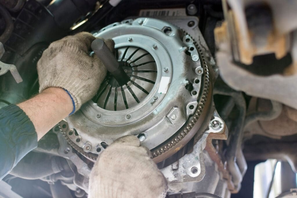 A mechanic wearing gloves skillfully installs or repairs a car clutch system, focusing on the pressure plate and clutch disc. In the automotive repair setting's backdrop, vehicle undercarriage parts hint at comprehensive car servicing alongside routine MOT testing sessions.