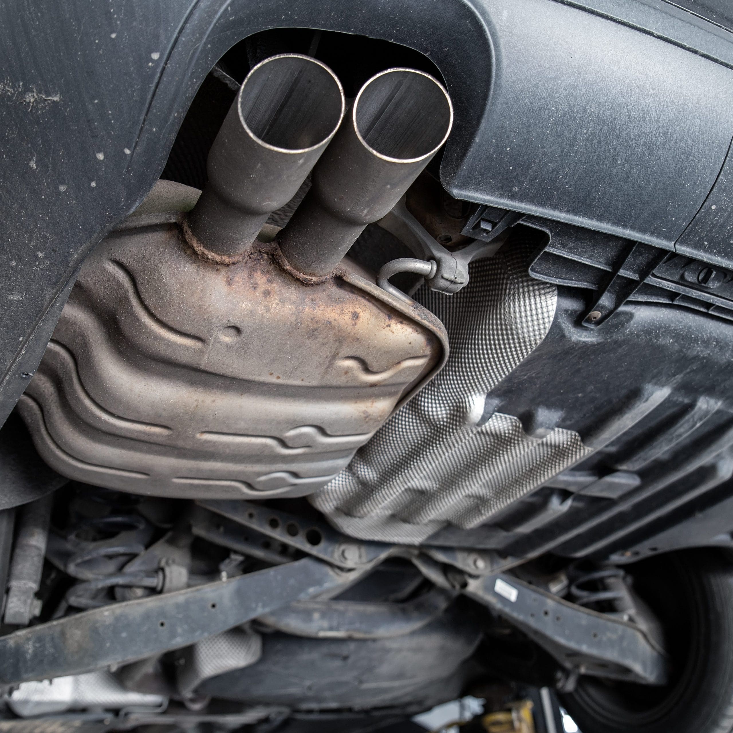 Underneath view of a vehicle shows a dual exhaust system and the muffler, hinting at recent checks by a skilled garage mechanic. The car's undercarriage looks slightly dirty, with visible exhaust pipes and metal components, alongside some protective shielding.