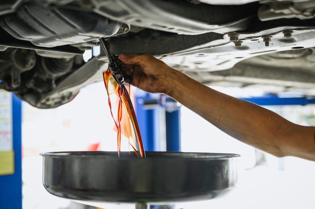 A person's hand drains dark motor oil from underneath a vehicle into a black container, highlighting the essential car servicing process. Set in an automotive workshop, the vehicle is raised on a lift, emphasizing routine maintenance alongside tasks like DPF cleaning and clutch inspection.