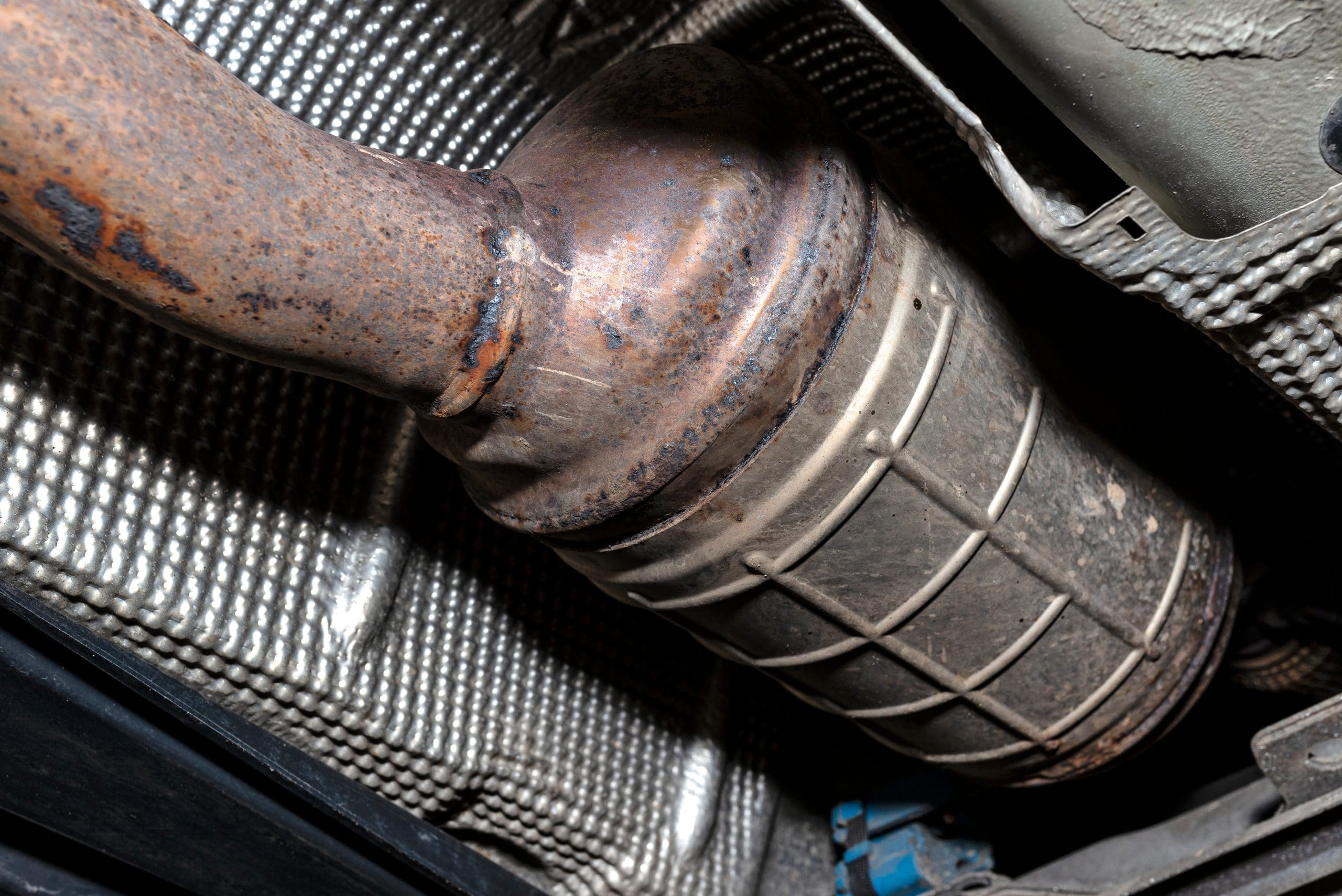 Close-up of an old, rusted catalytic converter under a vehicle. The metal surface is corroded, and it's attached to a textured protective heat shield. The surrounding area is part of the vehicle's undercarriage.